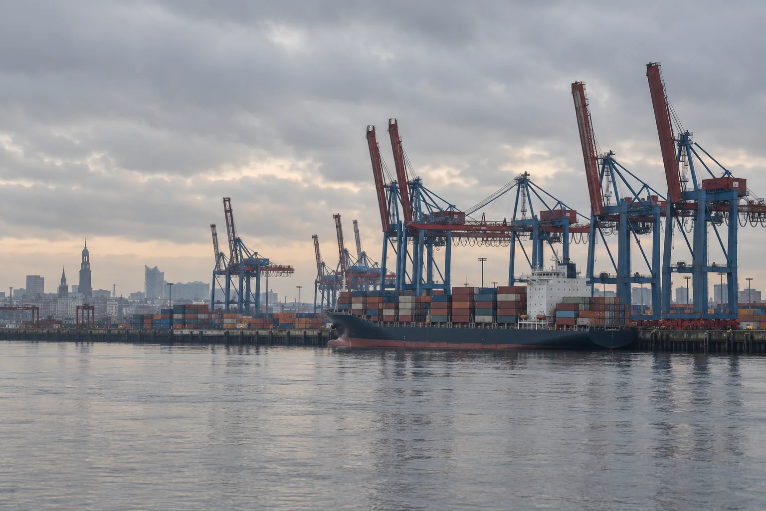 Stacked shipping containers and cranes at the port of Hamburg under a grey morning sky, seen from across the Elbe.